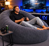 Man sitting on a bean bag chair playing video games in a room with gaming setup.